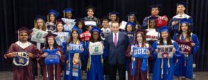 Group photo of 20 graduates in various colored cap and gowns posing with Coach Mike Krzyzewski on risers. Each graduate is holding a sweatshirt with their university name.